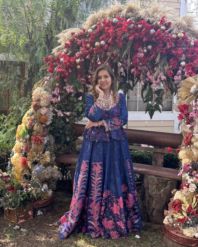Cris standing under an arch of flowers in a garden, wearing a long blue dress