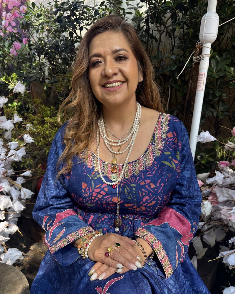 Cris sonriendo, sentada en un jardín con flores blancas, con vestido azul bordado y collares de perlas